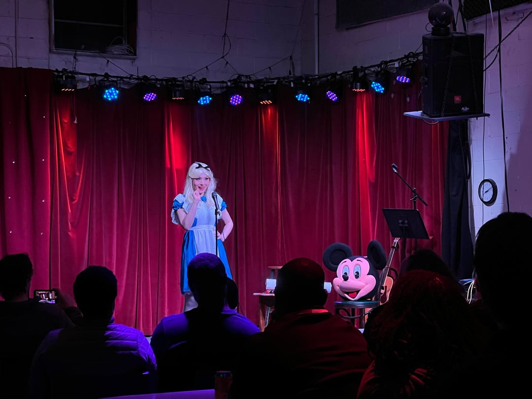 A comedian on stage wearing an Alice in Wonderland costume performs.