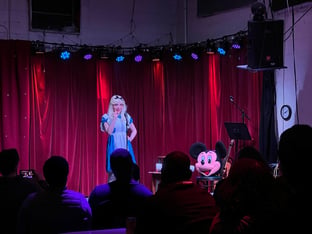 A comedian on stage wearing an Alice in Wonderland costume performs.