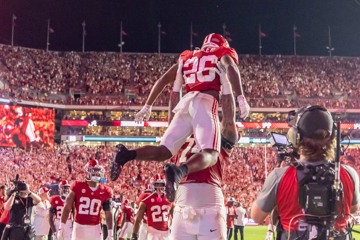 A football player raises another football player in the air in the middle of a crowded stadium.