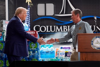Donald Trump, left, and Brian Kemp, right shake hands at press event, standing next to cases of water and boxes of diapers and other supplies.