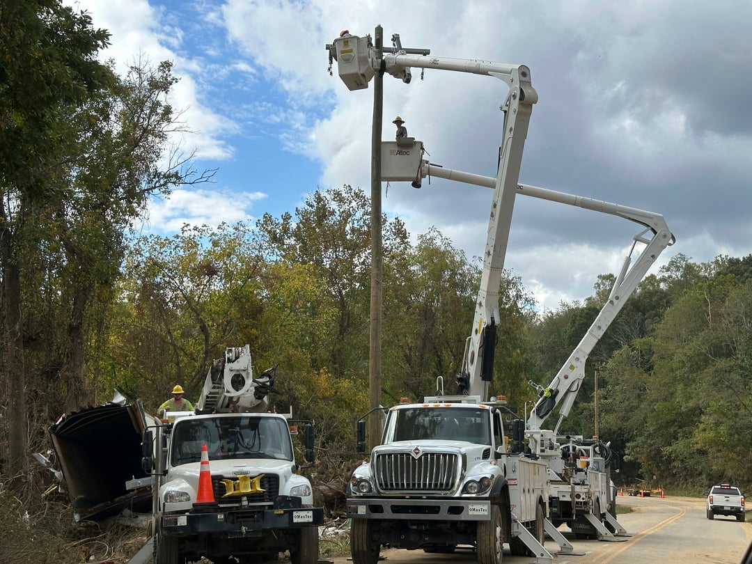 Several vehicles with long cranes are next to a site where there has been significant storm damage to trees.