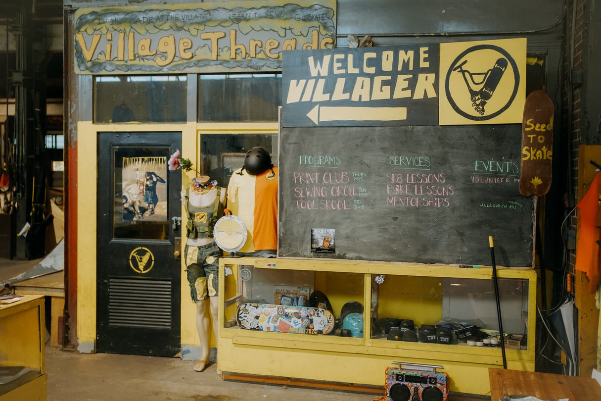 A small indoor storefront that says Village Threads, with a bulletin board in front of it that shows a variety of programming.