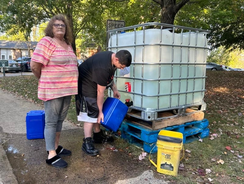 Teresa Thomas stands to the left as her son bends down to fill up buckets with water from a large tank.