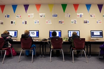 Students sitting at a row of desks and using desktop computers.