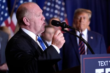 Tom Homan faces right, speaking, as President-elect Donald Trump looks on in the background.