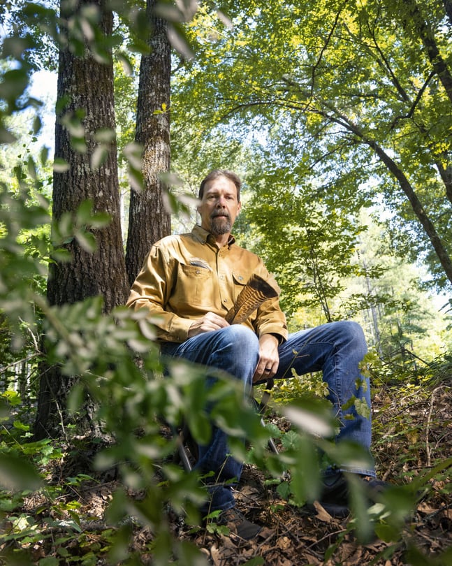 Mike Chamberlain in a yellow long sleeve shirt and jeans sits in the forest, surrounded by greenery, holding a wild turkey feather.