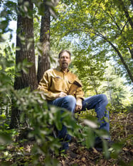 Mike Chamberlain in a yellow long sleeve shirt and jeans sits in the forest, surrounded by greenery, holding a wild turkey feather.