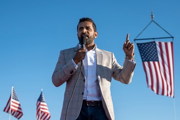 Kash Patel speaks into a microphone while standing in front of several American flags.