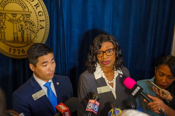 Democratic leaders Sam Park, Carolyn Hugley, and Tanya Miller stand in front of a blue curtain in front of a crowd of microphones.