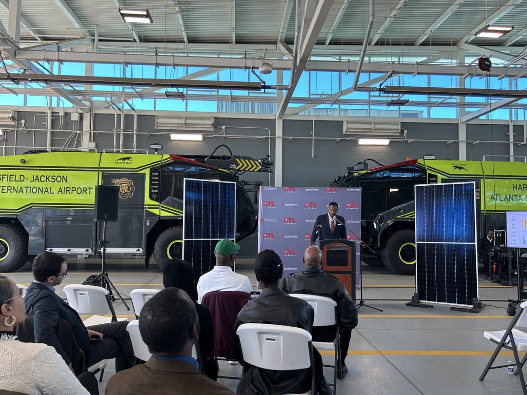 Mayor Andre Dickens stands behind a podium in front of two large green trucks. In front of him are a few rows of chairs in an arch, facing away from the photographer.