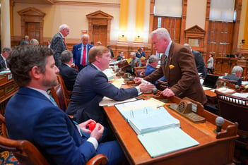 Republican State Sen. Blake Tillery and State Senate Majority Whip Randy Robertson speak on the floor of the Georgia State Senate in the Georgia State Capitol. Tillery is sitting in his seat and Robertson is standing in front of Tillery's desk.