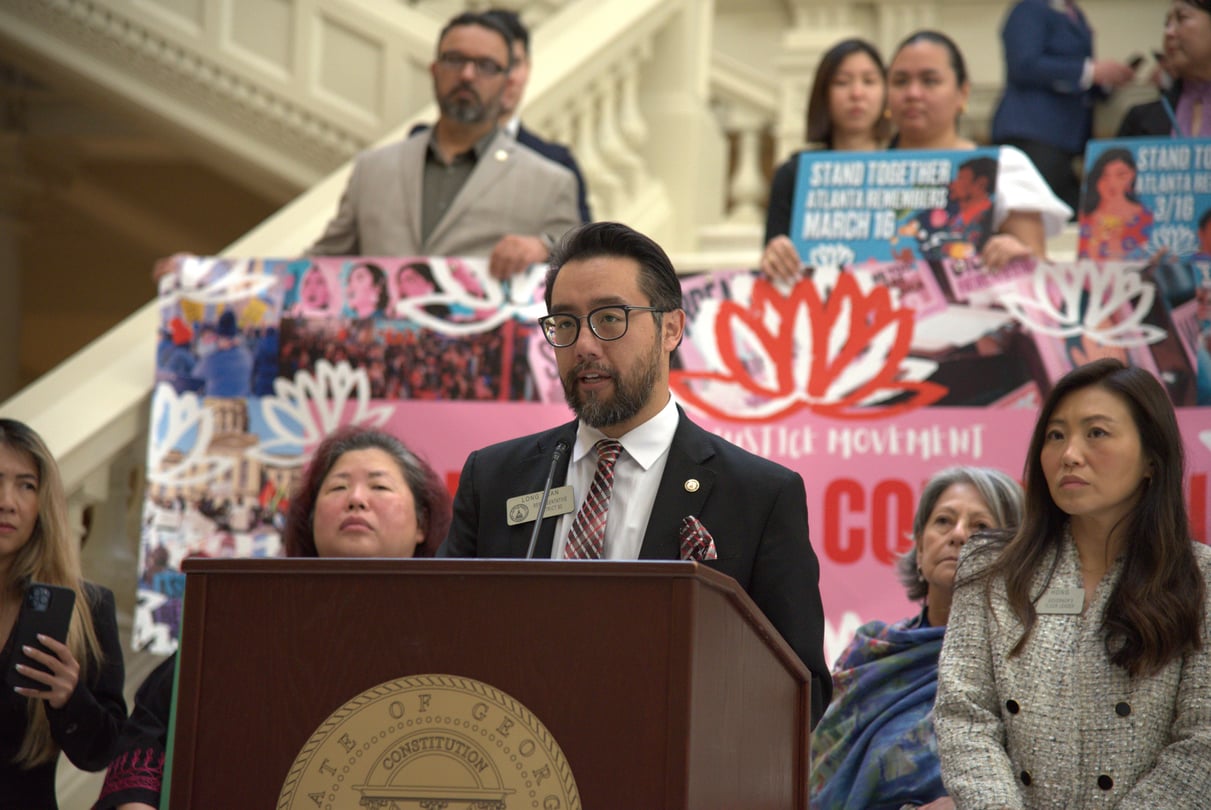 State Rep. Long Tran speaks at a podium, surrounded by lawmakers and activists holding signs relating to AAPI activism.