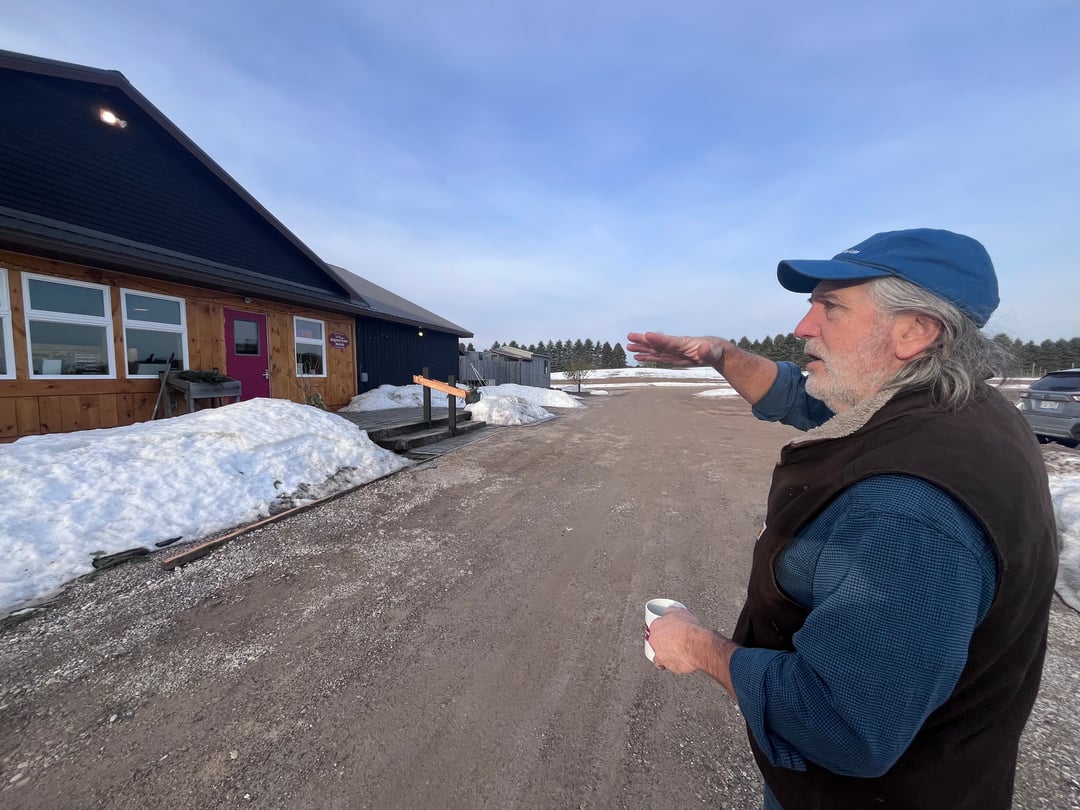 a bearded man gestures toward a metal-roofed building