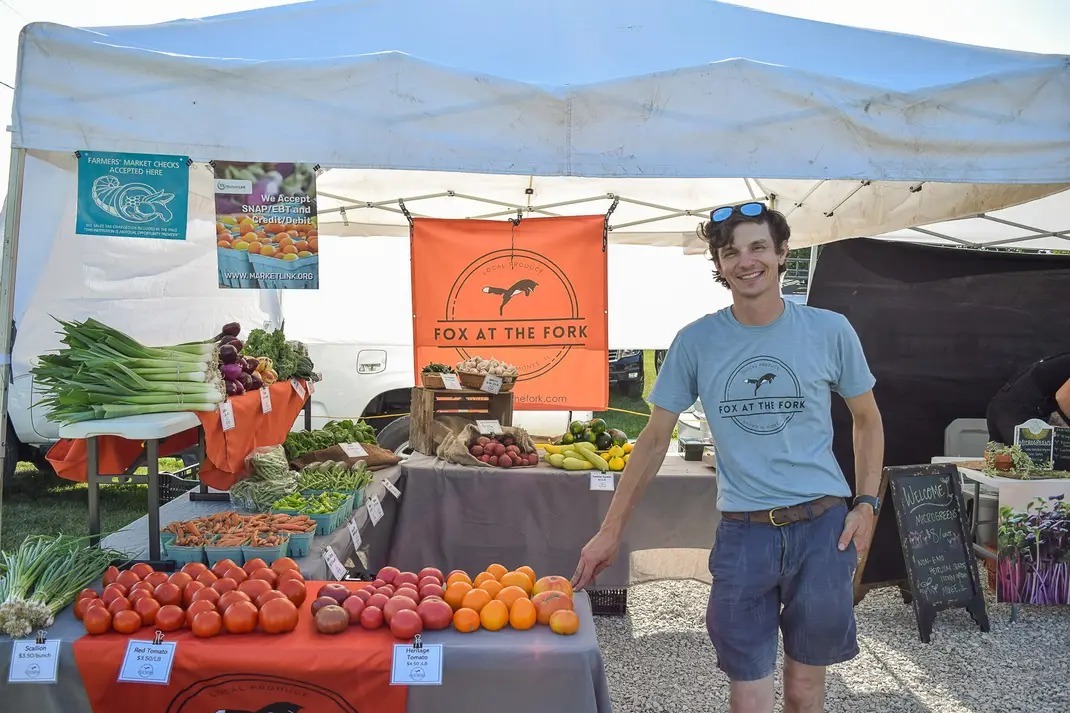 a man stands beside produce at a farmers market