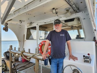 A man in a shirt and baseball cap stands on a fishing boat