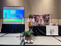 A woman stands behind a podium surrounded by soccer balls. The projector to the left of her shows on a screen saying FIFA World Cup 2025 Business Seminar.
