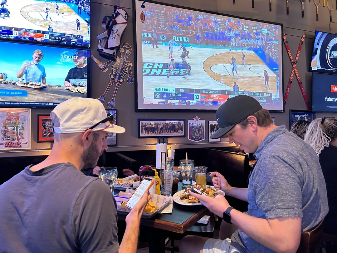 Two men with phones in their hands sit in front of a large screen showing a basketball game.