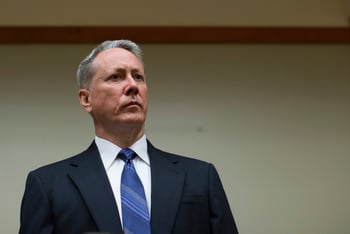A man in a suit and blue tie stands in front of a white wall.
