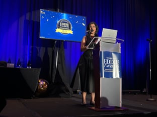 A brown woman with black hair wearing a black dress stands in front of a podium that says Eddie Awards 2025