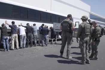 Employees stand in a line with their hands against a wall. Officers stand a few paces away from them.