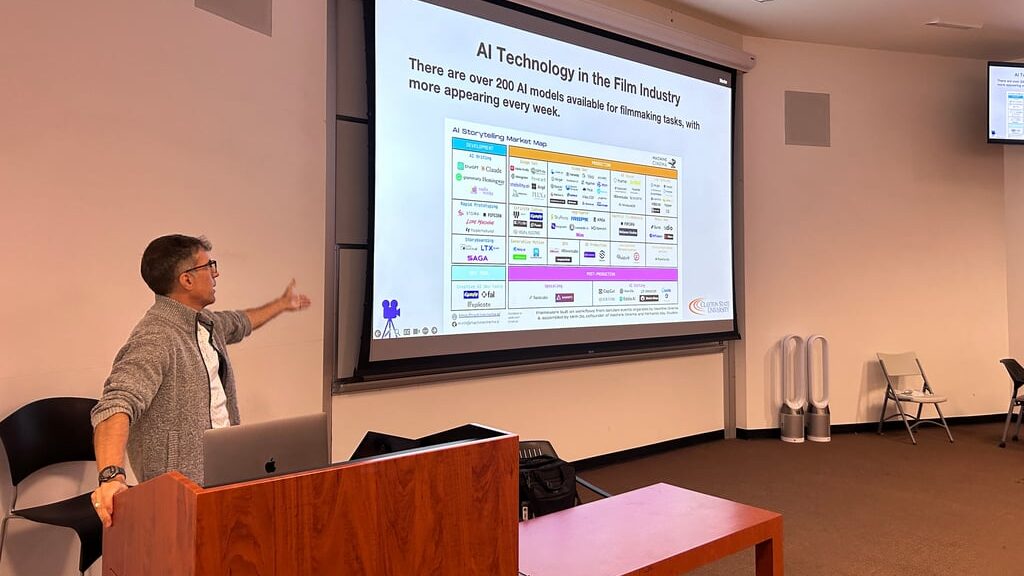A man teaching a class stands at a podium with a powerpoint behind him.