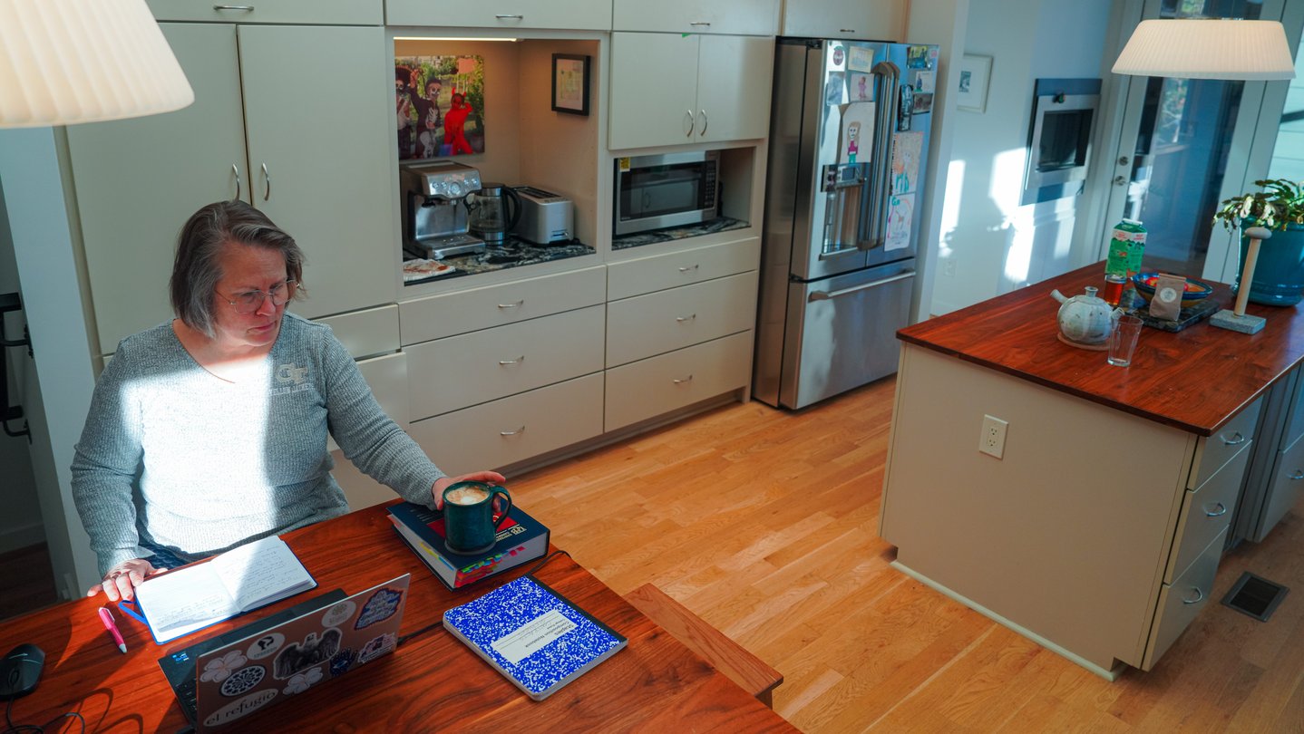 A woman sits at a desk in a kitchen watching an immigration hearing on a laptop screen.