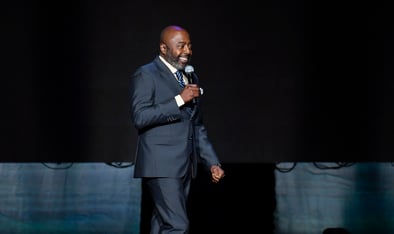 Comedian onstage in a dark suit, smiling as he holds a microphone