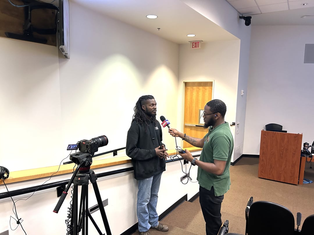 A man holds a mic up to another man in a classroom.
