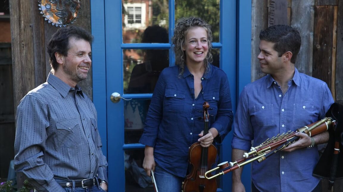 Trio smiling outside a blue door; fiddle and uilleann pipes visible.