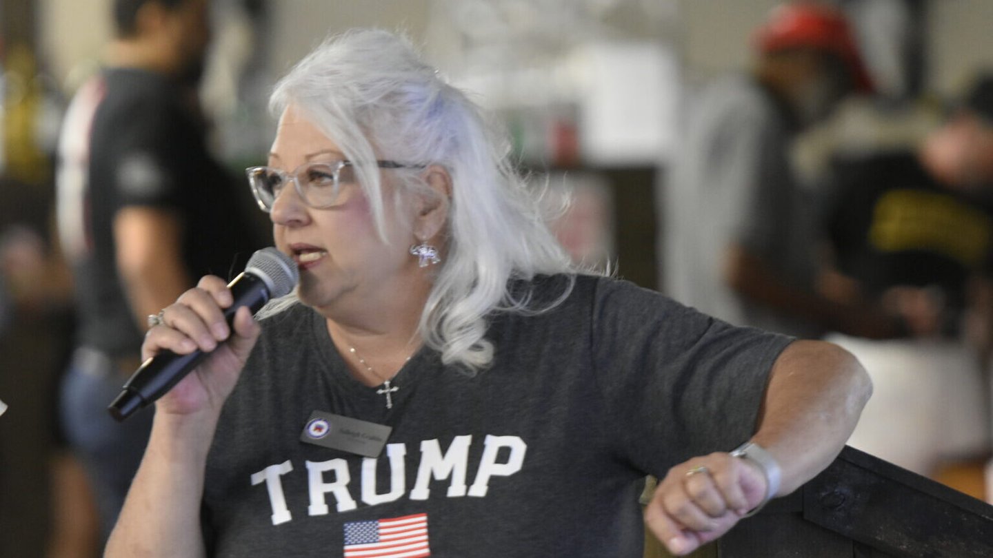 A woman with white hair and glasses, wearing a Trump shirt, speaks into a microphone