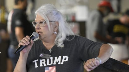 A woman with white hair and glasses, wearing a Trump shirt, speaks into a microphone