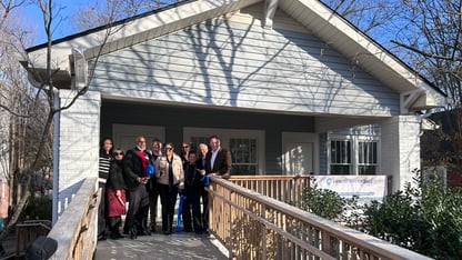A group of people stand in front of a house holding cut pieces of ribbon.