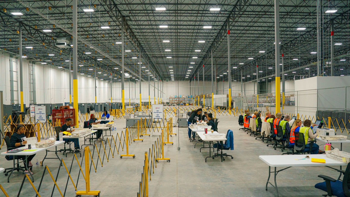 The interior of a large building is shown with rows of tables and workers preparing to process election ballots