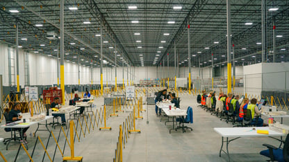 The interior of a large building is shown with rows of tables and workers preparing to process election ballots