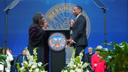 A woman and a man stand facing each other at a podium against a blue background for a ceremony.