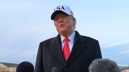 President Donald Trump talks in front of microphones while wearing a white hat with USA on it