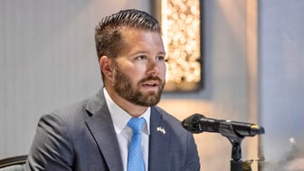 A man wearing a light blue tie and a grey suit sitting behind a microphone.