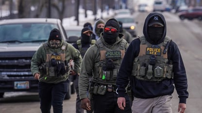 Immigration and Customs Enforcement officers walk down a street with cars behind them