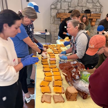 Volunteers stand at a long table with a yellow tablecloth assembling a long time of open-faced cheese sandwiches.