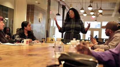 A group of people sit around a table and listen while a person standing talks