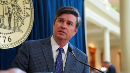 Matt Brass stands in front of a blue background for a press conference and speaks into a microphone and a cell phone.