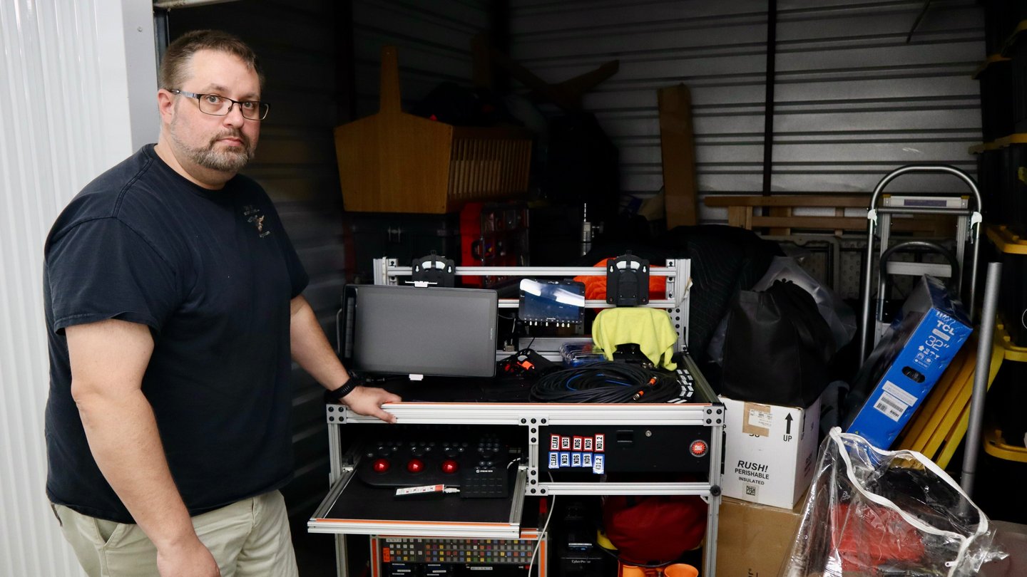 A man in a black t-shirt stands by a bunch of film equipment in a storage unit