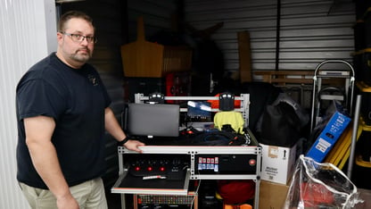 A man in a black t-shirt stands by a bunch of film equipment in a storage unit