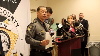 A sheriff stands and speaks at a podium for a press conference while other people watch