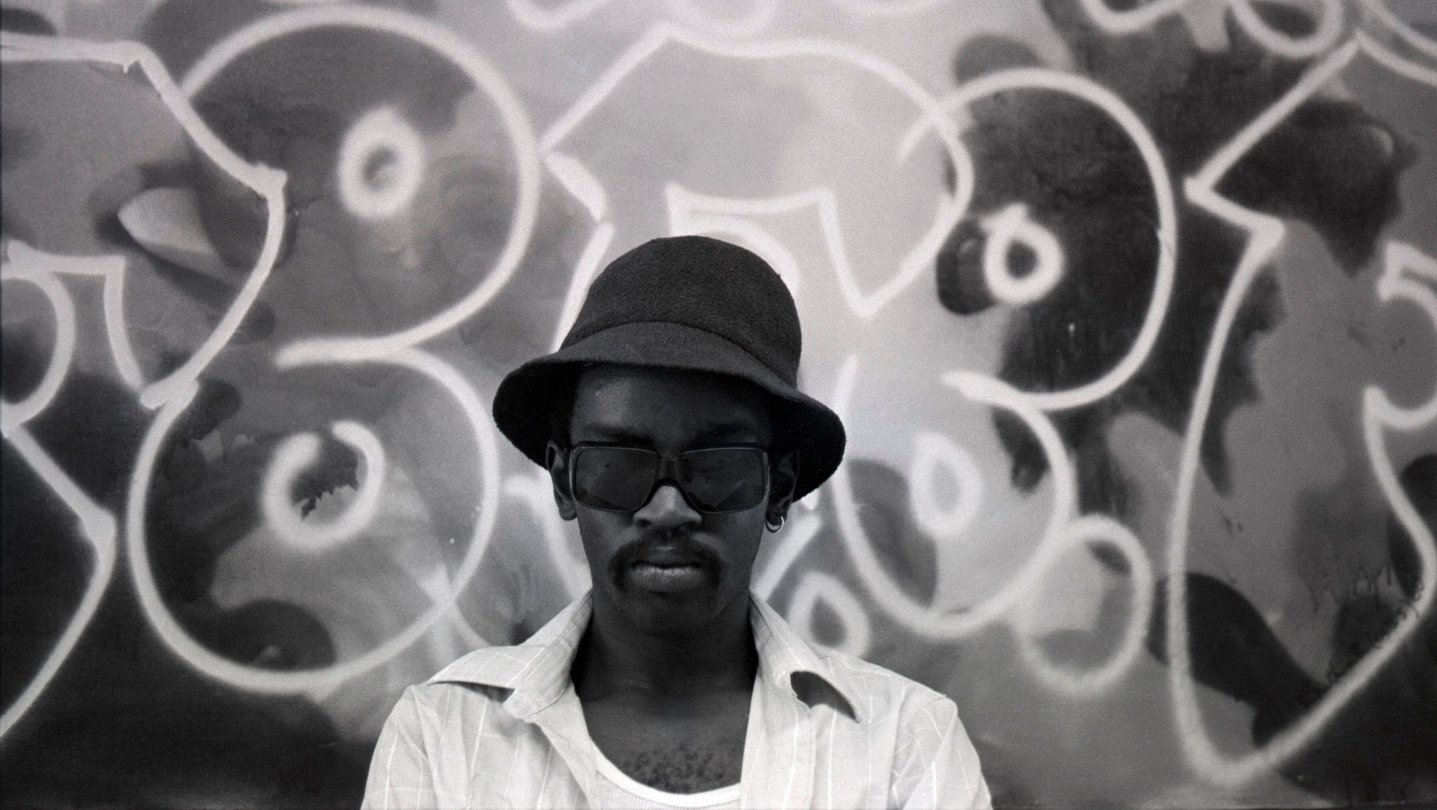 Fab Five Freddy stands before a graffiti mural in a black and white portrait, wearing a bucket hat and dark glasses.