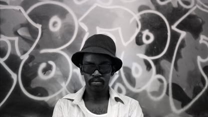 Fab Five Freddy stands before a graffiti mural in a black and white portrait, wearing a bucket hat and dark glasses.