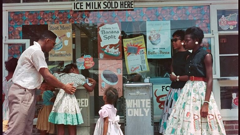 A photograph of several people outside of a glass window of an ice milk store, drinking from a water fountain labeled Colored Only