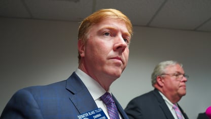 A closeup shot of a lawmaker with another lawmaker behind him in a room with a white wall.
