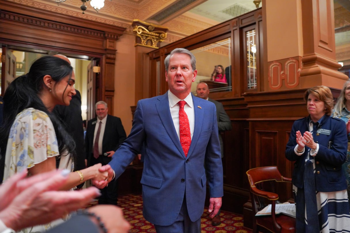 Brian Kemp, wearing a blue suit and a red tie, walks into the House chamber surrounding by people applauding him