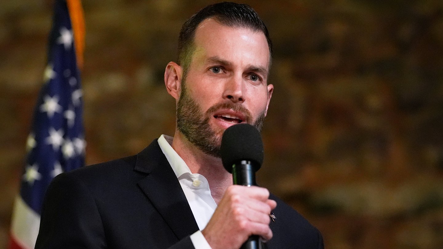 Clay Fuller speaks into a microphone in front of an American flag